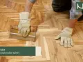 Ręce w rękawiczkach pracują nad drewnianą posadzką, układając parkiet w jodełkę.