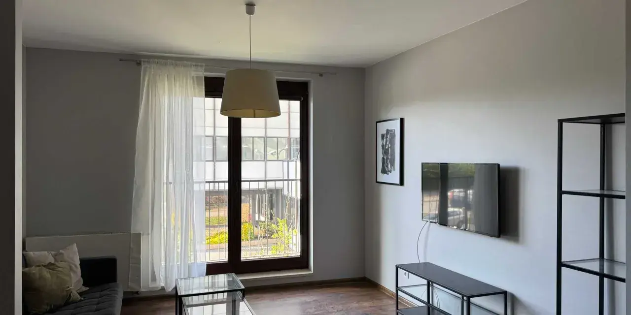 Nowoczesny salon z szarą kanapą, szklanymi stolikami i telewizorem. Widok z okna na zielony dziedziniec, idealne miejsce na relaks w śródmieście Wrocław.
