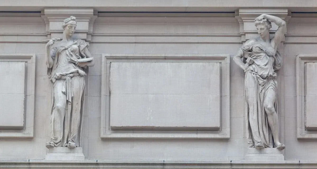 Dwie kamienne rzeźby architektoniczne w antycznym stylu, przedstawiające kobiety w togach, zdobią fasadę budynku.