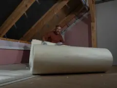 Mężczyzna w rękawiczkach przygotowuje ocieplenie dachu, rozwijając dużą rolkę izolacji.