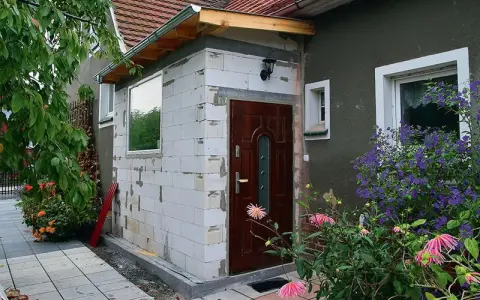 Trwa rozbudowa budynku gospodarczego. Nowa część z białych pustaków, brązowe drzwi, okno i zielone rośliny.