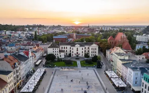 Zachód słońca nad rynkiem, gdzie widać piękną pierzeję kamienic i ratusz. Ludzie spacerują po placu, ciesząc się ciepłym wieczorem.
