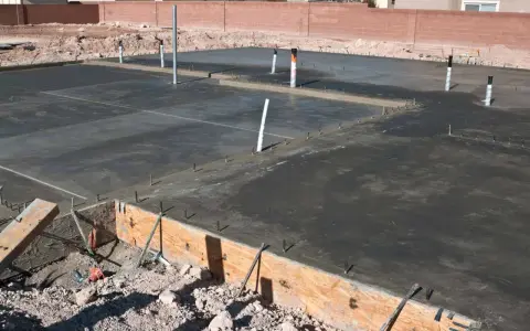 Świeżo wylany beton na płycie fundamentowej, widoczne rury i zbrojenie. Widać przygotowania do izolacji pionowej fundamentów.