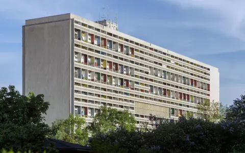 Długi budynek mieszkalny w stylu modernizm, z rzędami balkonów w kolorowych obramowaniach, otoczony zielenią.