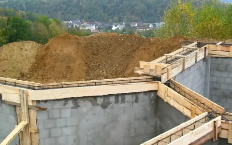 Fundamenty domu w budowie, zbrojenie i szalunki. Widać kopce ziemi i zabudowania w tle. Ważne jest pozwolenie na budowę, by wszystko szło zgodnie z planem.