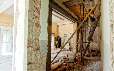 Trwa częściowe wyburzenie ściany nośnej. Widać robotników, rusztowania i gruz.