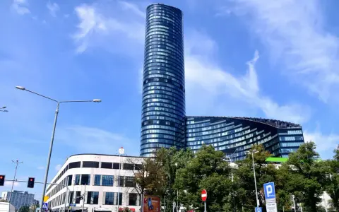 Wysoki, szklany budynek Sky Tower we Wrocławiu, z pewnością ma wiele pięter. Widać też niższy budynek i drzewa.