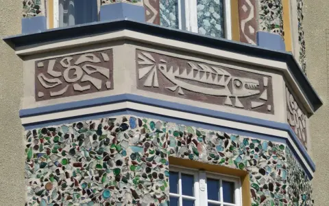 Sgraffito na fasadzie budynku: stylizowana ryba i abstrakcyjne wzory. Dolna część ozdobiona mozaiką z kolorowego szkła.