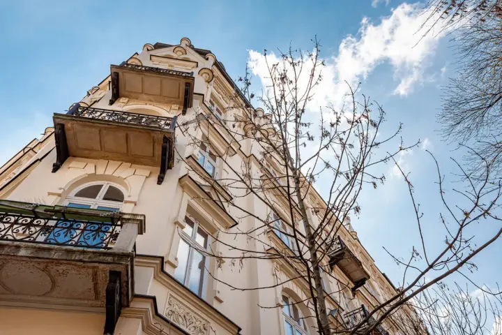 Elegancka kamienica z balkonami i ozdobnymi detalami, widziana z dołu na tle błękitnego nieba z chmurami.