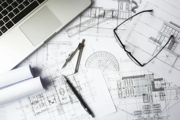 Na biurku architekta leżą plany budowlane, laptop, okulary i cyrkiel. Trwają prace nad przedmiarem robót.