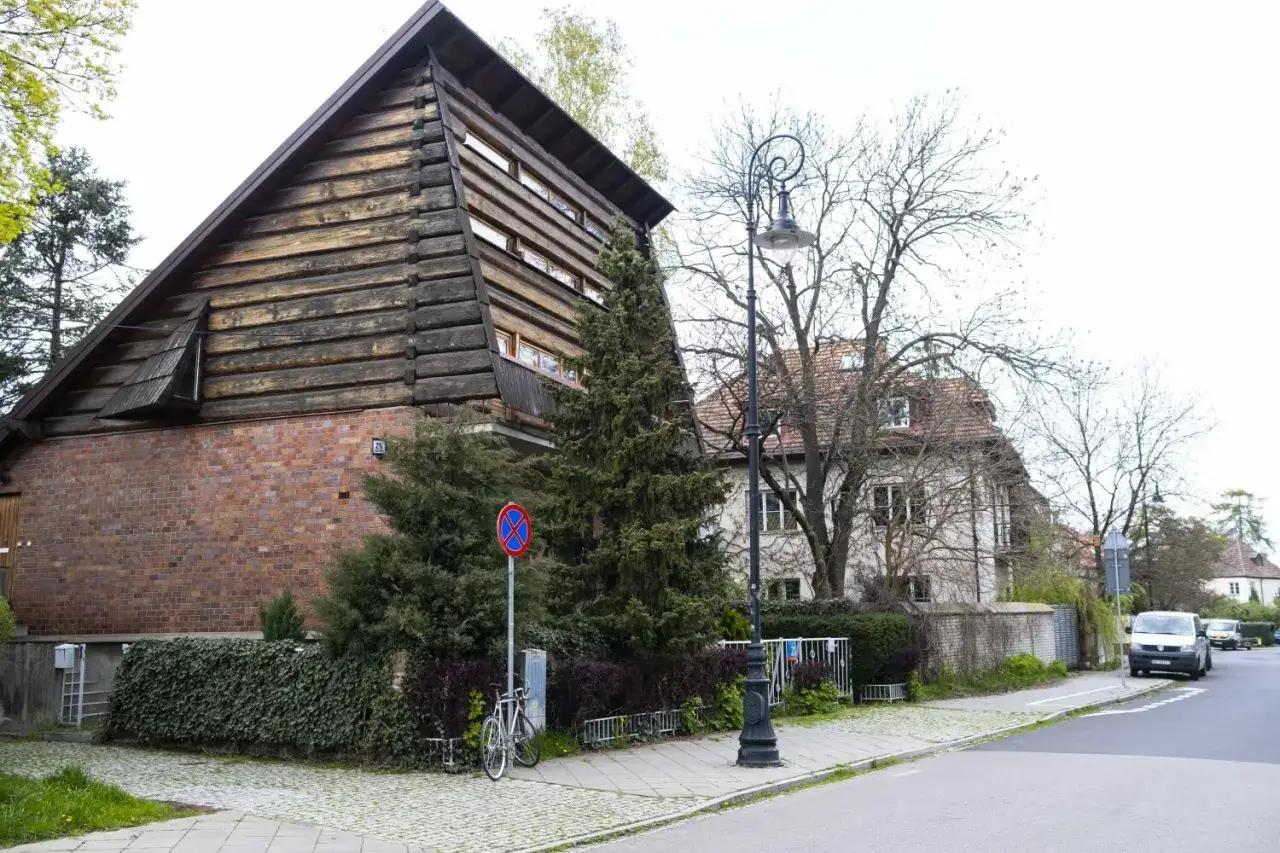 Drewniany dom z ceglaną podstawą i charakterystycznym dachem, stojący przy ulicy na Żoliborzu Oficerskim. Obok rower i zabytkowa latarnia.