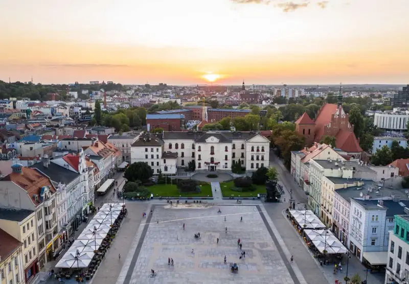 Zachód słońca nad rynkiem, gdzie widać piękną pierzeję kamienic i ratusz. Ludzie spacerują po placu, ciesząc się ciepłym wieczorem.